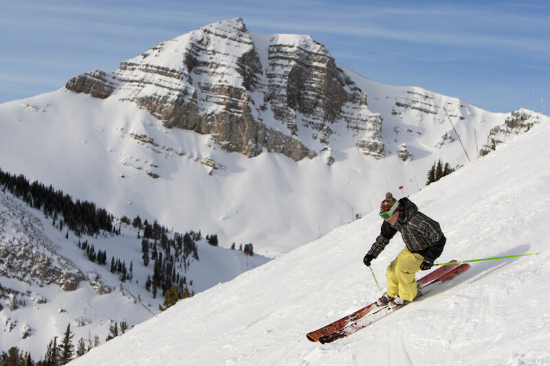 A skier descends a snowy slope with majestic, rugged mountains in the background on a clear day.